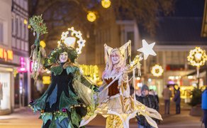 Zwei Personen in fantasievollen Kostümen mit Lichterketten, eine trägt einen Sternstab, die andere hält einen Mistelzweig, im Hintergrund weihnachtlich beleuchtete Straße mit Menschen.