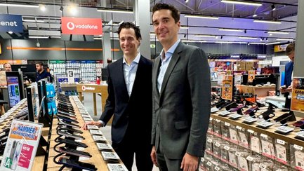 Zwei Männer stehen lächelnd nebeneinander in einem Elektronikmarkt. Im Vordergrund und Hintergrund sind verschiedene ausgestellte Elektrogeräte zu sehen.