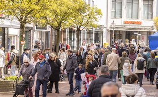 Belebte Fußgängerzone mit vielen Menschen unterschiedlichen Alters, die zwischen Geschäften flanieren; einige schieben Kinderwagen oder Gehhilfen, während Bäume und Ladenfronten die Straße säumen.