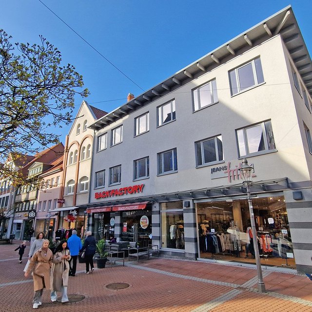 Mehrstöckiges Geschäftshaus an einer belebten Fußgängerzone mit mehreren Passanten und Bäumen.