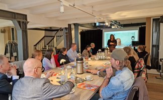 Menschen sitzen an einem langen Tisch im Peiner „Schwan“ mit sichtbaren Stahlträgern und hören Peine-Marketing-Geschäftsführerin Anja Barlen-Herbig zu, die vor einer Leinwand spricht.