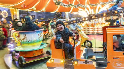 Kind sitzt in orangefarbenem Spielzeug-LKW auf Karussell mit bunten Lichtern und weiteren Fahrgeschäften im Hintergrund.