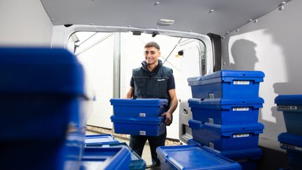 Ein Mann trägt blaue Transportboxen aus einem Lieferfahrzeug.