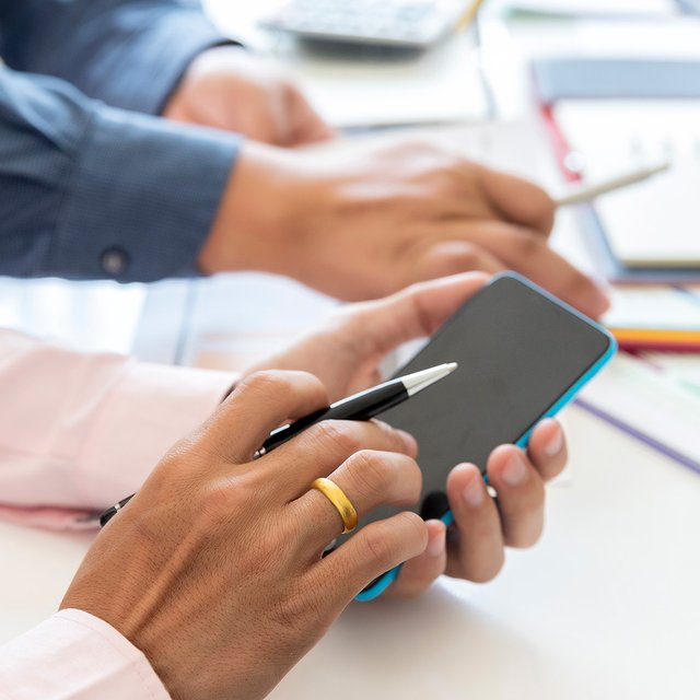 Hände von zwei Personen, eine hält ein Smartphone, die andere zeigt mit einem Stift darauf, im Hintergrund Schreibtisch mit Papieren und Laptop.