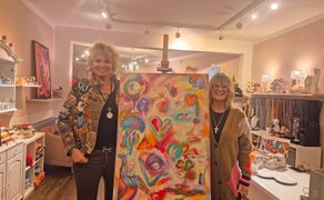 Zwei Personen stehen in einem Raum mit Kunstwerken, zwischen ihnen eine Staffelei mit einem farbenfrohen abstrakten Gemälde.