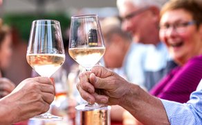 Zwei Personen stoßen mit Weingläsern an, im Hintergrund unscharfe Menschen bei einer Veranstaltung im Freien.