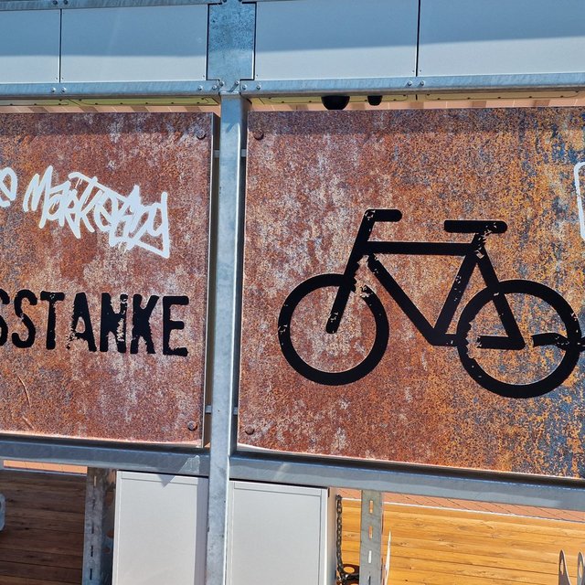 Rostfarbene Metalltafeln an einer Fahrradstation mit der Aufschrift „Peiner Stahlrosstanke“, einem Fahrradsymbol mit Stecker und dem Hinweis „Gratis laden / Free charge“.