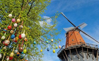 Windmühle mit rot gedecktem Kegel und hölzernem umlaufenden Balkon neben Baumzweigen mit zahlreichen bunt bemalten Ostereiern an Bändern.