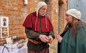 Zwei Personen in historischer Kleidung stehen vor einer Backsteinwand und betrachten gemeinsam alte Werkzeuge und Anschauungsobjekte auf einem Tisch; der Mann erklärt etwas, während die Frau aufmerksam zuhört.