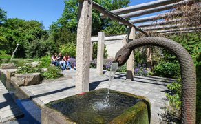 Ein steinerner Brunnen mit einem geschwungenen Wasserauslauf steht im Vordergrund. Dahinter befinden sich eine Pergola, blühende Beete, Bäume und drei sitzende Personen in einer gepflegten Parkanlage.