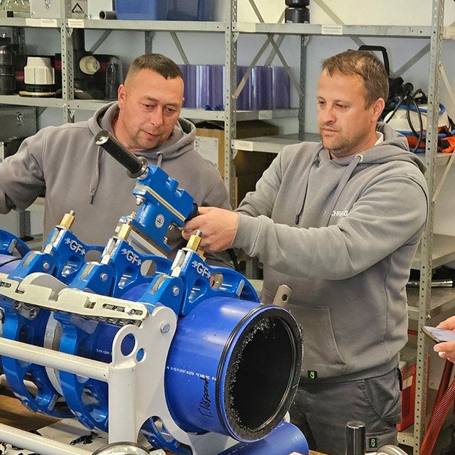 Vier Männer stehen in einer Werkstatt an einer technischen Apparatur, in die ein Kunststoffrohr eingespannt ist.