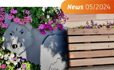 Das Bild zeigt eine Bank mit einem stilisierten Wolf dahinter und bunten Blumen. 