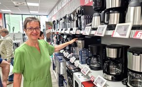 Person zeigt in einem Elektronikfachmarkt auf verschiedene Kaffeemaschinen und Küchengeräte, die in einem Verkaufsregal präsentiert werden.