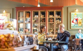 Zwei Personen sitzen an einem Tisch in einem Café, trinken aus Tassen und unterhalten sich.