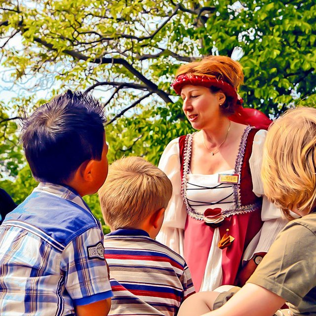 Eine Gruppe von Kindern steht im Freien unter grünen Bäumen und hört aufmerksam einer Frau zu, die eine historische Stadtführung durchführt. Die Frau trägt ein mittelalterlich wirkendes Kleid mit Kopftuch und spricht lebendig zu den Kindern, die sich neugierig um sie versammelt haben. Die Szene wirkt freundlich und lebendig bei sonnigem Wetter.