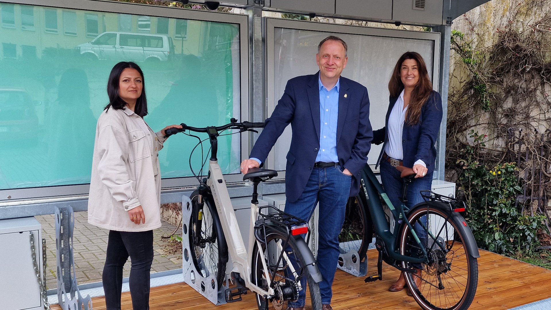 Das Bild zeigt drei Menschen, die mit Fahrrädern an einem BikePort stehen. 