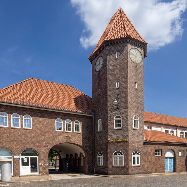 Ein großes Backsteingebäude mit rotem Ziegeldach und markantem Uhrturm steht an einem Platz. Der Turm hat auf mehreren Seiten Zifferblätter. Das Gebäude verfügt über Rundbögen im Eingangsbereich und mehrere Fensterreihen. Im Vordergrund ist eine gepflasterte Fläche zu sehen, rechts und links stehen einzelne Bäume. Der Himmel ist blau mit wenigen Wolken.