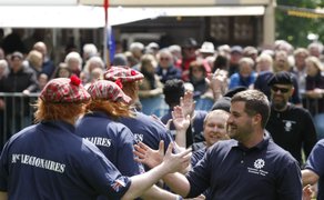Teilnehmer in schottischer Tradition kleiden sich in Tartanmützen und Teamshirts und klatschen sich jubelnd ab, während im Hintergrund zahlreiche Zuschauer das Geschehen bei einem Highland-Gathering verfolgen.