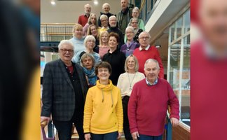 Gruppe von Menschen unterschiedlichen Alters in einem Innenraum mit Treppe und Geländer im Hintergrund.