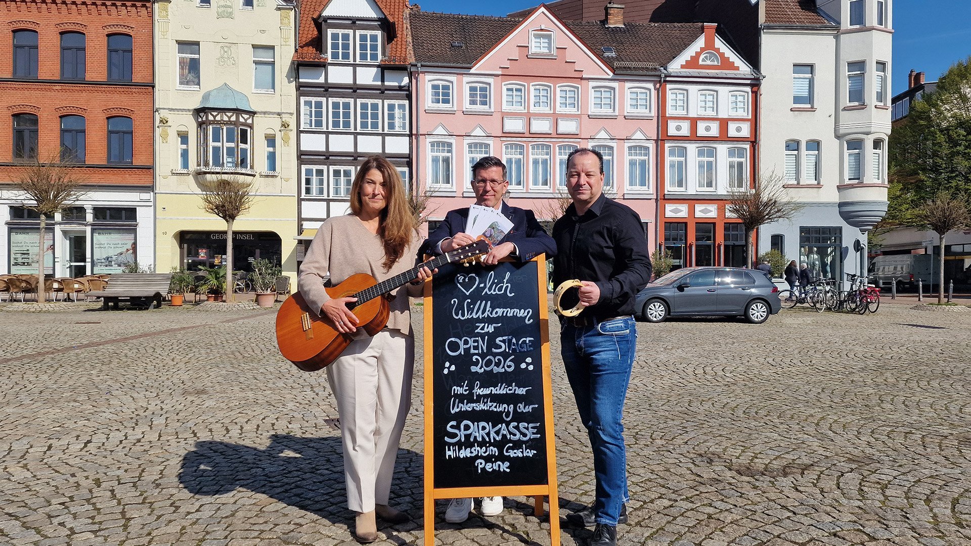 Drei Personen stehen auf dem Historischen Marktplatz in Peine vor bunten Fachwerkhäusern; eine Frau hält eine Gitarre, ein Mann ein Tamburin, in der Mitte steht eine Tafel mit der Aufschrift „Herzlich Willkommen zur Open Stage 2026“, die auf das Musik- und Kulturangebot im Freien hinweist.