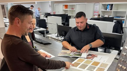 Eine Frau und ein Mann stehen an einem Tressen mit Mustern und Katalogen, die ein Mann auf der anderen Seite des Tresens präsentiert. Im Hintergrund Regale und Arbeitslätze mit Computern.