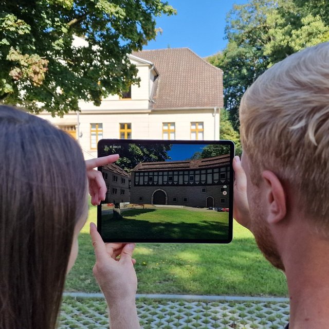 Zwei Personen betrachten auf einem Tablet eine digitale Rekonstruktion eines historischen Gebäudes im Park vor einem heutigen Bauwerk.