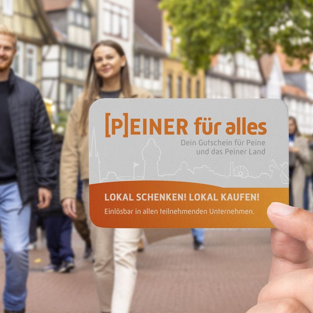 Eine Hand hält den Gutschein „[P]EINER für alles“ in der Peiner Fußgängerzone, im Hintergrund gehen Passanten durch die Innenstadt.