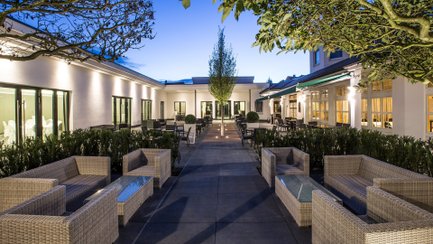 Innenhof eines Hotels mit Sitzgruppen aus Rattanmöbeln und Bäumen entlang eines gepflasterten Wegs bei Dämmerung.