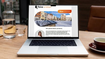 Laptop auf Holztisch zeigt Newsletter mit Überschrift „Peine erleben“ und einem Foto eines historischen Marktplatzes.