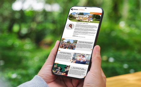 Eine Person hält ein Smartphone in der Hand. Auf dem Display ist der digitale Newsletter „Peine erleben“ geöffnet, der mit Bildern und Beiträgen zu Veranstaltungen, Freizeitangeboten und Erlebnissen in Peine gefüllt ist. Im Hintergrund ist eine unscharfe grüne Parklandschaft zu sehen.