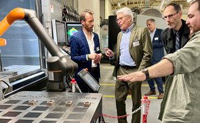Mehrere Männer in Geschäftskleidung stehen in einer Werkhalle vor einem Industrieroboter mit orangefarbenem Arm und Steuerpult.