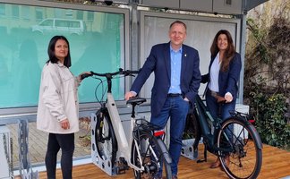 Das Bild zeigt drei Menschen, die mit Fahrrädern an einem BikePort stehen. 