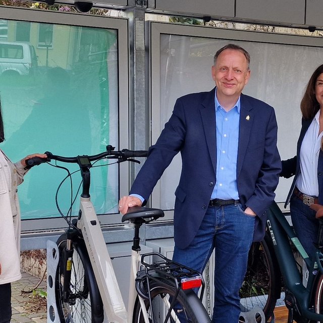 Das Bild zeigt drei Menschen, die mit Fahrrädern an einem BikePort stehen. 