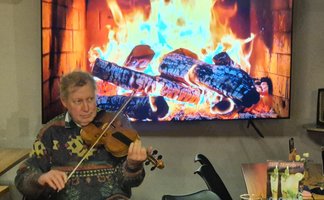 Das Bild zeigt einen Musiker, der Geige spielt. Im Hintergrund wird ein Lagerfeuer auf einem Fernsehgerät gezeigt. 