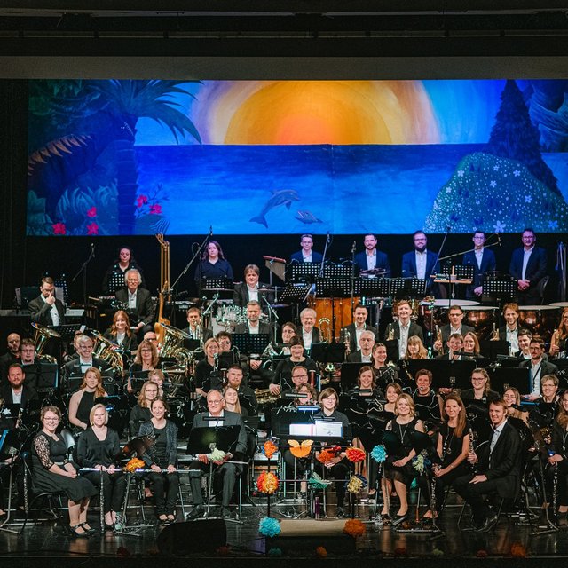 Das Bild zeigt das Stadtorchester Peine auf einer Bühne mit einem blauen Bühnenbild im Hintergrund. 