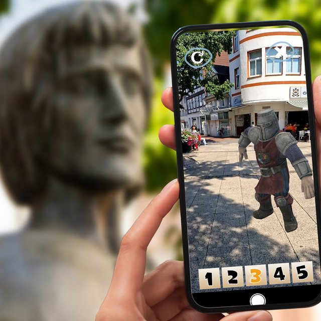 Das Bild zeigt im Vordergrund Hände, die ein Handy halten, auf dem ein Comic-Ritter zu sehen ist. Im Hintergrund sieht amn die Skulptur von Graf Gunzelin.