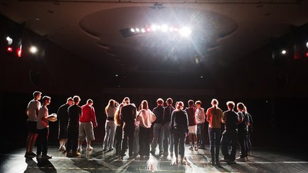 Das Bild zeigt eine Gruppe von Menschen auf einer Theaterbühne, die mit dem Rücken zum Fotografen stehen. 