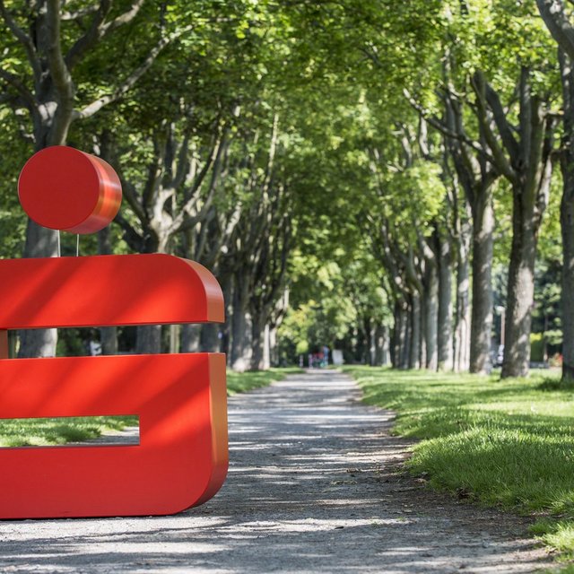 Das Bild zeigt das Logo der Sparkasse auf einem Weg in einem Park. 
