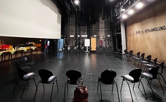 Das Bild zeigt eine Theaterbühne, auf der sich zahlreiche leere Stühle befinden, die im Kreis aufgestellt sind. 