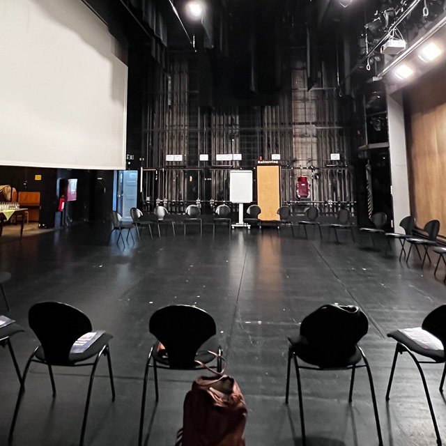 Das Bild zeigt eine Theaterbühne, auf der sich zahlreiche leere Stühle befinden, die im Kreis aufgestellt sind. 