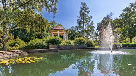 Das Bild zeigt den Teich im Stadtpark Peine mit dem Pavillon im Hintergrund. 