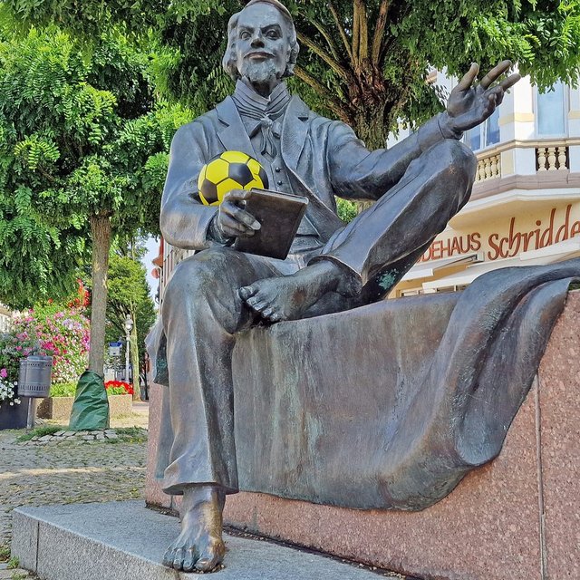 Das Bild zeigt die Skulptur von Friedrich von Bodenstedt auf dem Historischen Marktplatz, der einen gelben Fußbal in der Hand trägt.