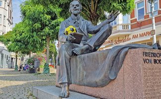 Das Bild zeigt die Skulptur von Friedrich von Bodenstedt auf dem Historischen Marktplatz, der einen gelben Fußbal in der Hand trägt.
