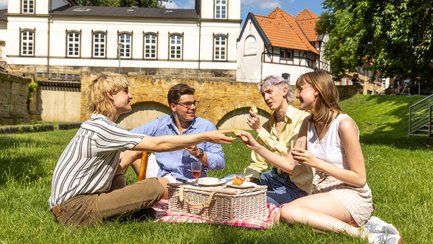 Das Bild zeigt eine Gruppe von vier jungen Leuten, die im Burgerpark auf dem Rasen sitzen und ein Picknick veranstalten. 
